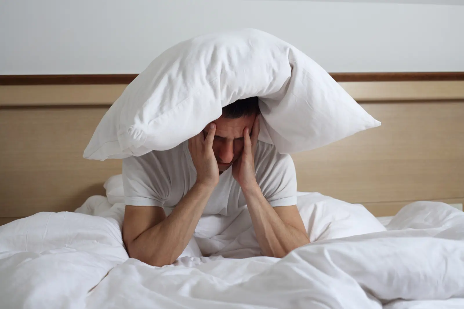 Homme fatigué assis sur un lit, avec un oreiller sur la tête, souffrant de troubles du sommeil et d’insomnie.